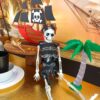 a skeleton sits leaning against a wooden cake stand with a pirate ship on top. He sits next to a wooden palm tree