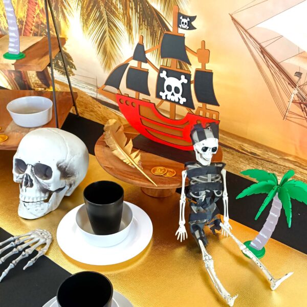 a skeleton sits leaning against a wooden cake stand with a pirate ship on top. He sits next to a wooden palm tree