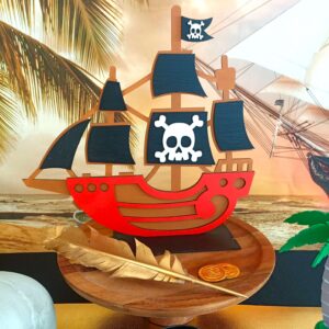 A wooden pirate ship sits a top a wooden cake stand. There is a golden quill and gold coins sitting in front of it